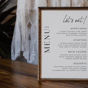 Op de afbeelding: Een ingelijst menu met de tekst "let's eat!" bovenaan. Het menu bevat voorgerechten, soepen, hoofdgerechten en desserts. Het menu ligt op een houten tafel.