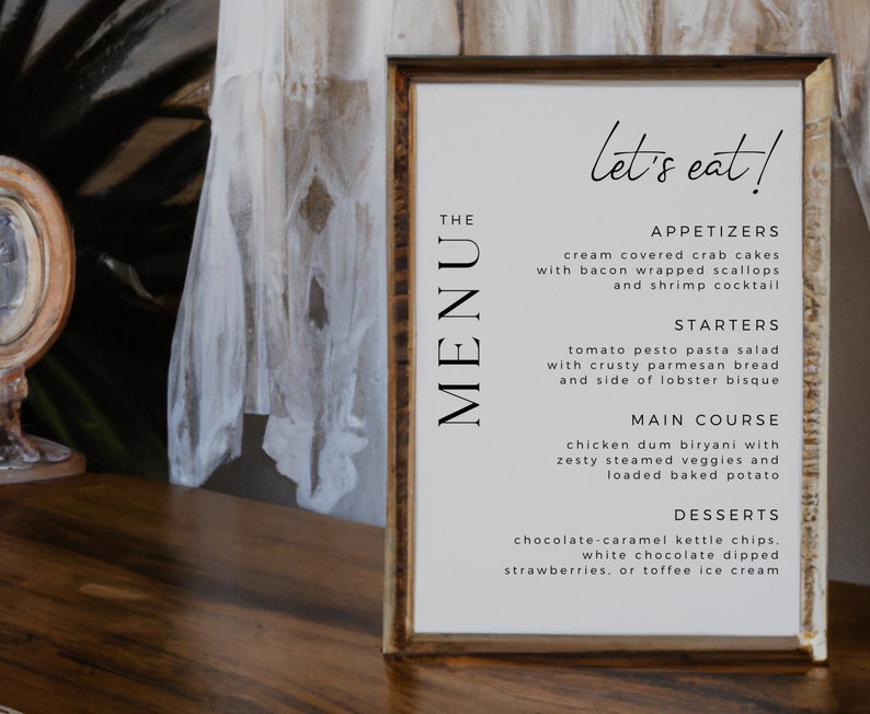 Op de afbeelding: Een ingelijst menu met de tekst "let's eat!" bovenaan. Het menu bevat voorgerechten, soepen, hoofdgerechten en desserts. Het menu ligt op een houten tafel.