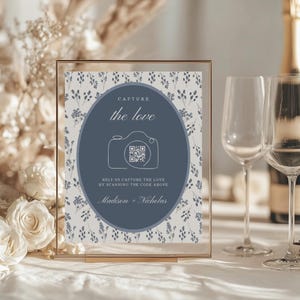 May include: A clear framed sign with a floral background and a navy blue oval in the center. The oval has a camera graphic and the words "Capture the love". Below the oval is the text "Help us capture the love by scanning the code above" and the names "Madison + Nicholas".