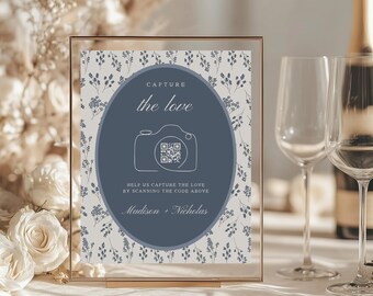 May include: A clear framed sign with a floral background and a navy blue oval in the center. The oval has a camera graphic and the words "Capture the love". Below the oval is the text "Help us capture the love by scanning the code above" and the names "Madison + Nicholas".