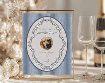 May include: A framed sign with a blue striped background and an oval design. The oval contains a photo of a golden retriever and the text "PLEASE TAKE A pawty treat FUR MY FRIENDS Lilly". The frame is gold.