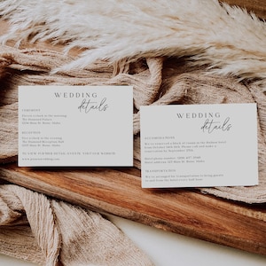 May include: Two white cards with black text detailing wedding ceremony and reception information, including the time, location, and website. The cards are on a wooden surface with a beige fabric overlay.