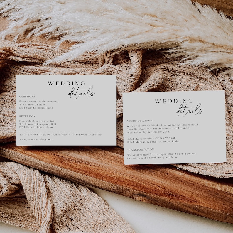 May include: Two white cards with black text detailing wedding ceremony and reception information, including the time, location, and website. The cards are on a wooden surface with a beige fabric overlay.