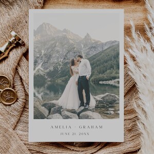 May include: A wedding announcement card with a photo of a couple kissing in front of a mountain lake. The text on the card reads "Amelia & Graham, June 21, 20XX".