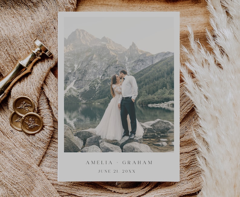May include: A wedding announcement card with a photo of a couple kissing in front of a mountain lake. The text on the card reads "Amelia & Graham, June 21, 20XX".