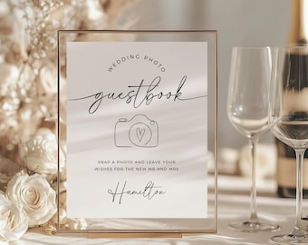 May include: A white sign with black text that reads "Wedding Photo Guestbook" with a simple line drawing of a camera with a heart inside. The sign also says "Snap a photo and leave your wishes for the new Mr. and Mrs. Hamilton".