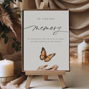 May include: A white sign with a gold frame on a wooden easel. The sign reads "In Loving Memory" with a handwritten script font. Below the text is a butterfly with brown and orange wings flying towards an open hand. The text below the butterfly reads "Of those who can't be with us today but are forever in our hearts."