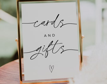 May include: A gold-framed sign with the words "cards and gifts" in elegant black script, with a small heart at the bottom. The sign is displayed on a wooden surface, suitable for weddings or special events.