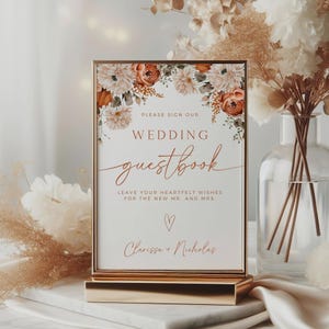 May include: A framed wedding guestbook sign with floral accents in shades of orange, white, and green. The sign reads "PLEASE SIGN OUR WEDDING guestbook" and includes space for names. The sign is displayed on a gold stand.