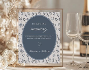 May include: A framed sign with the text "IN LOVING memory" in a blue oval, surrounded by floral patterns. The sign includes the names "Madison + Nicholas" and a heart symbol. The frame is gold, and the background includes champagne flutes and flowers.