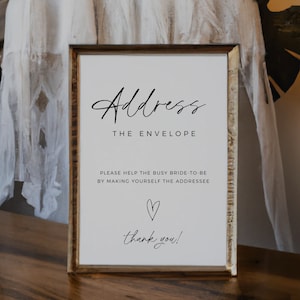 May include: A white sign with a black frame and the text "Address the Envelope" in a cursive font. The sign also includes the text "Please help the busy bride-to-be by making yourself the addressee" and a heart symbol. The sign ends with the text "thank you!"