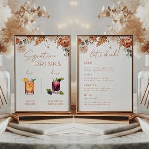 May include: Two framed signs with gold trim and bases. The left sign reads "Signature Drinks" with "his" and "hers" below. The right sign says "let's drink!" with drink options. Floral accents adorn the top.