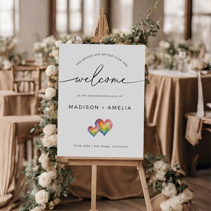 May include: White welcome sign with the text "Two brides are better than one" and "welcome to the bachelorette party of MADISON + AMELIA 10.25.20XX | San Diego, California". The sign features two watercolor hearts in rainbow colors.