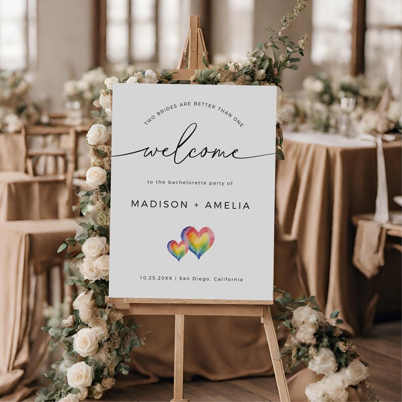 May include: White welcome sign with the text "Two brides are better than one" and "welcome to the bachelorette party of MADISON + AMELIA 10.25.20XX | San Diego, California". The sign features two watercolor hearts in rainbow colors.