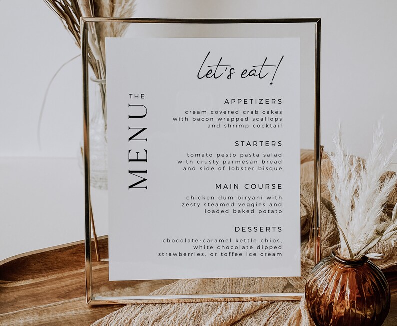 Op de afbeelding: Een wit menu bord met een gouden frame, met een lijst van voorgerechten, starters, hoofdgerecht en desserts. Het menu is getiteld "The Menu" en heeft de tekst "let's eat!" bovenaan.