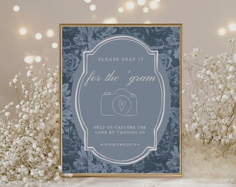 May include: A framed sign with a blue floral background and a central oval design. The oval contains a camera graphic with a heart, and the text "PLEASE SNAP IT for the 'gram". The sign encourages guests to tag the wedding.