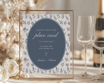 May include: A framed place card with a floral pattern and a navy blue oval in the center. The text on the card reads "PLEASE TAKE YOUR place card TO YOUR SEAT so we know what you're having to eat." The names "Madison + Nicholas" are at the bottom.