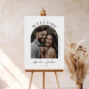 Op de afbeelding: Een wit welkomsbord met een zwarte boog en een foto van een stel. De tekst luidt "Welkom op de bruiloft van Amelia + Graham 06.21.2028".