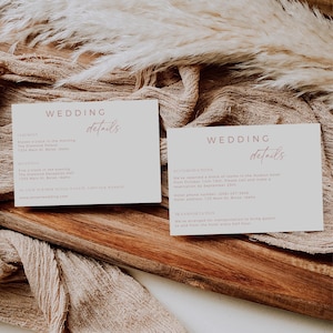 May include: Wedding details card with a pink script font. The card includes information about the ceremony, reception, accommodations, and transportation. The card is on a wooden surface with a beige fabric and a dried grass plume.