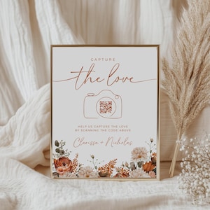 May include: A framed sign with the text "CAPTURE the love" in a handwritten font, with a camera graphic and a QR code. The sign also includes the names "Clarissa + Nicholas" and floral accents in shades of orange, white, and brown.