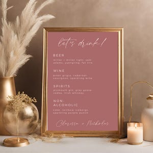 Könnte beinhalten: Ein rosa Schild mit goldener Umrandung, auf dem "let's drink!" steht und verschiedene Arten von alkoholischen und alkoholfreien Getränken aufgelistet sind. Das Schild steht auf einem Tisch mit einer goldenen Vase und einer Kerze.