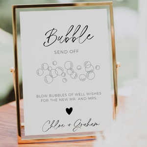 Op de afbeelding: Een wit bord met zwarte tekst en een zwart hart in een gouden lijst. Het bord zegt "Bubble Send Off" en "Blaas bellen vol goede wensen voor het nieuwe echtpaar Chloe + Graham".
