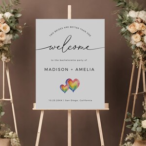 May include: A welcome sign for a bachelorette party with the text "Two brides are better than one" and "Welcome to the bachelorette party of Madison + Amelia". The sign also includes the date "10.25.20XX" and location "San Diego, California". The sign features two watercolor hearts in rainbow colors.
