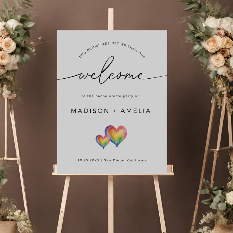 May include: A welcome sign for a bachelorette party with the text "Two brides are better than one" and "Welcome to the bachelorette party of Madison + Amelia". The sign also includes the date "10.25.20XX" and location "San Diego, California". The sign features two watercolor hearts in rainbow colors.
