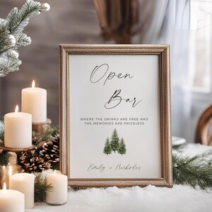 May include: A gold-framed sign with the text "Open Bar" and the phrase "Where the drinks are free and the memories are priceless". The sign also includes a small illustration of three pine trees and the names "Emily + Nicholas".