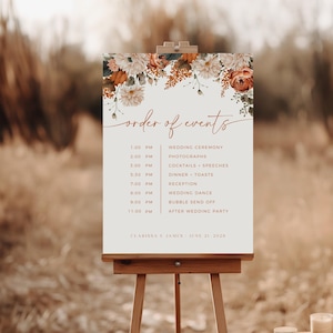 May include: A wooden easel displays a wedding order of events sign. The sign features a floral design with orange and cream-colored flowers. The text includes the wedding schedule, names, and date.