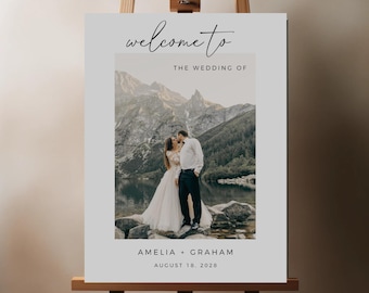 May include: White welcome sign with a photo of a couple kissing in front of a mountain lake. The text reads "Welcome to the Wedding of Amelia + Graham August 18, 2028".