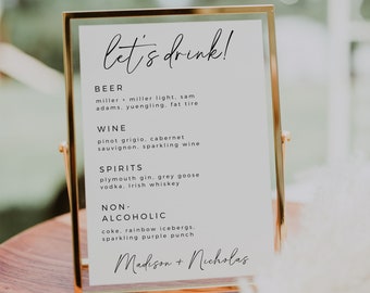 May include: A white sign with a gold frame lists the drinks available at a wedding reception. The sign reads "let's drink!" and lists beer, wine, spirits, and non-alcoholic options. The sign is signed "Madison + Nicholas".