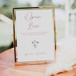 Pode incluir: Uma placa com moldura dourada, fundo branco e texto rosa que diz "Open Bar" com o slogan "Where the drinks are free and the memories are endless". A placa também apresenta uma pequena ilustração floral e os nomes "Amelia + Nicholas" na parte inferior.