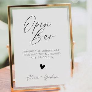 May include: A white sign with black calligraphy that reads "Open Bar" and the text "Where the drinks are free and the memories are priceless". Below the text is a small black heart and the names "Olivia + Graham". The sign is in a gold frame.
