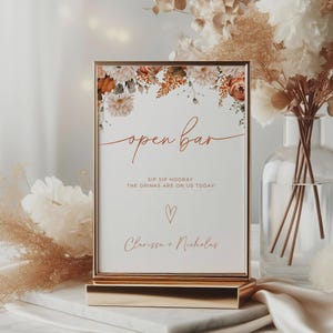 Puede incluir: Un letrero con marco dorado con las palabras "open bar" en escritura elegante, adornado con detalles florales en tonos melocotón y blanco. El letrero dice "SIP SIP HOORAY THE DRINKS ARE ON US TODAY!" con un corazón y nombres debajo.