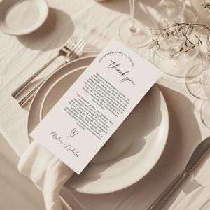 Könnte beinhalten: Eine Hochzeitstischdekoration mit einer weißen Tischkarte mit der Aufschrift "Thank you" in eleganter Schrift. Die Karte liegt auf einer weißen Serviette, mit Gabel, Teller und Gläsern. Die Karte enthält auch die Namen "Madison + Nicholas".