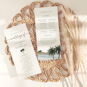 Può includere: Due biglietti bianchi per il programma del matrimonio con testo nero, con i nomi "Clarissa e Nicholas" e una scena di spiaggia tropicale. I biglietti sono su un sottobicchiere intrecciato con erba della pampa essiccata.
