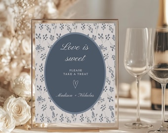 May include: A framed sign with the text "Love is sweet, Please take a treat" in a blue oval, with the names "Madison + Nicholas" below. The background has a floral pattern. The frame is gold.