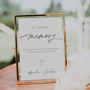 May include: A white sign with a gold frame, reading "In loving memory of those who can't be with us today but are forever in our hearts." A heart symbol and the names "Amelia + Graham" are also included.