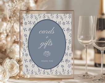 May include: A gold-framed sign with a blue oval that reads "cards + gifts" and "thank you." The background is white with a floral pattern. Two wine glasses and a champagne bottle are visible in the background.
