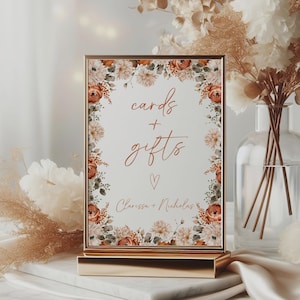 May include: A framed sign with the words "cards + gifts" in elegant script, surrounded by a floral border of peach and cream-colored flowers. The sign is displayed on a gold stand, perfect for a wedding or special event.