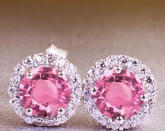 May include: A pair of pink gemstone stud earrings with a halo of small, clear stones. The earrings are set in a silver metal and are displayed on a light gray surface. The gemstones are a vibrant pink color.