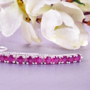 May include: A silver bracelet featuring a line of oval-cut, bright pink gemstones. The bracelet has a fine chain and a sliding clasp for adjustable sizing. White flowers and foliage are in the background.