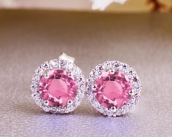 May include: A pair of stud earrings featuring a round, pink gemstone surrounded by a halo of smaller, clear stones. The earrings are set in a silver-colored metal and rest on a light gray surface. The background includes white floral accents.