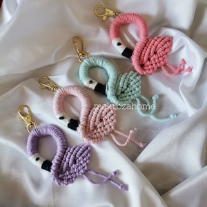 May include: Five macrame flamingo keyrings in shades of pink, light blue, lavender, and peach. Each flamingo has a gold-coloured clasp and a black and white beak. The keyrings are displayed on a white, silky fabric.