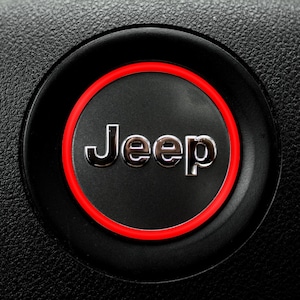 May include: Close-up of a black steering wheel with a circular emblem. The emblem features the silver word "Jeep" on a black background, encircled by a thin red ring. The steering wheel has a textured surface.