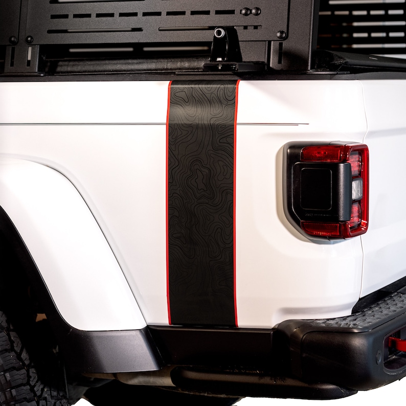 K&ouml;nnte beinhalten: Ein wei&szlig;er Pick-up mit einem schwarzen und roten Streifen an der Seite. Der Streifen zeigt ein topografisches Kartenmuster.