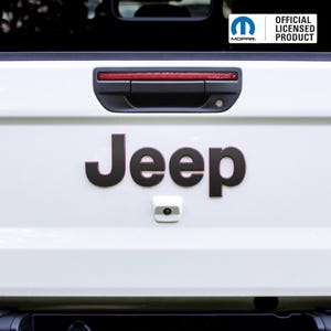 May include: White vehicle tailgate with black "Jeep" lettering and a backup camera. The Mopar logo and "OFFICIAL LICENSED PRODUCT" text are visible. A red light bar is above the handle.