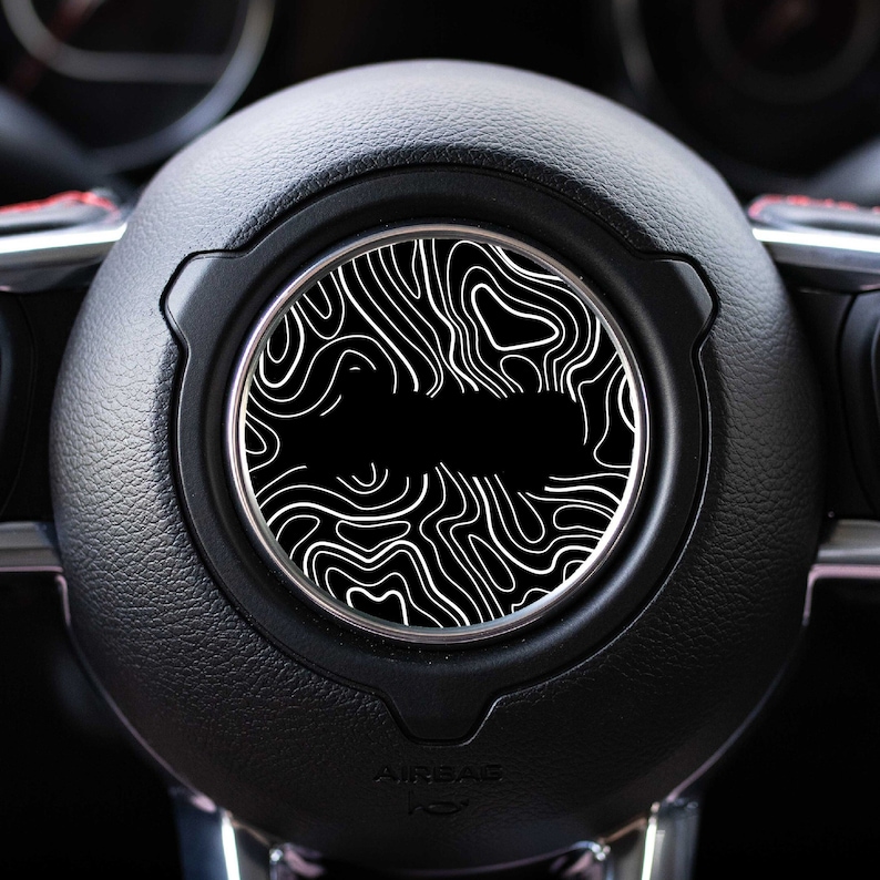 May include: A black leather car steering wheel with a white patterned design in the center. The design is a series of swirling lines and curves.