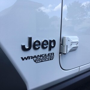May include: Close-up of a white Jeep Wrangler Unlimited Sport. The Jeep logo is in silver, with the Wrangler Unlimited Sport text below it in black. The door hinge is visible on the right side.